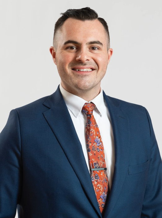 A man in a navy suit and orange tie smiling towards the camera.